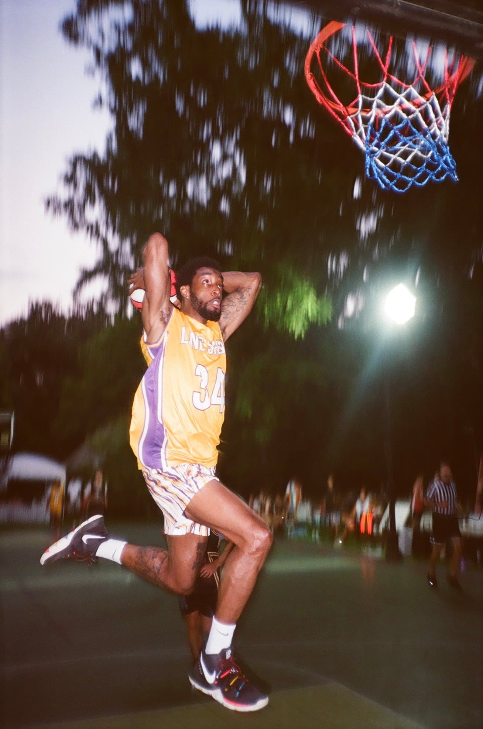 Player in a yellow and purple number 34 jersey rising for a dunk at dusk, red, white and blue net above