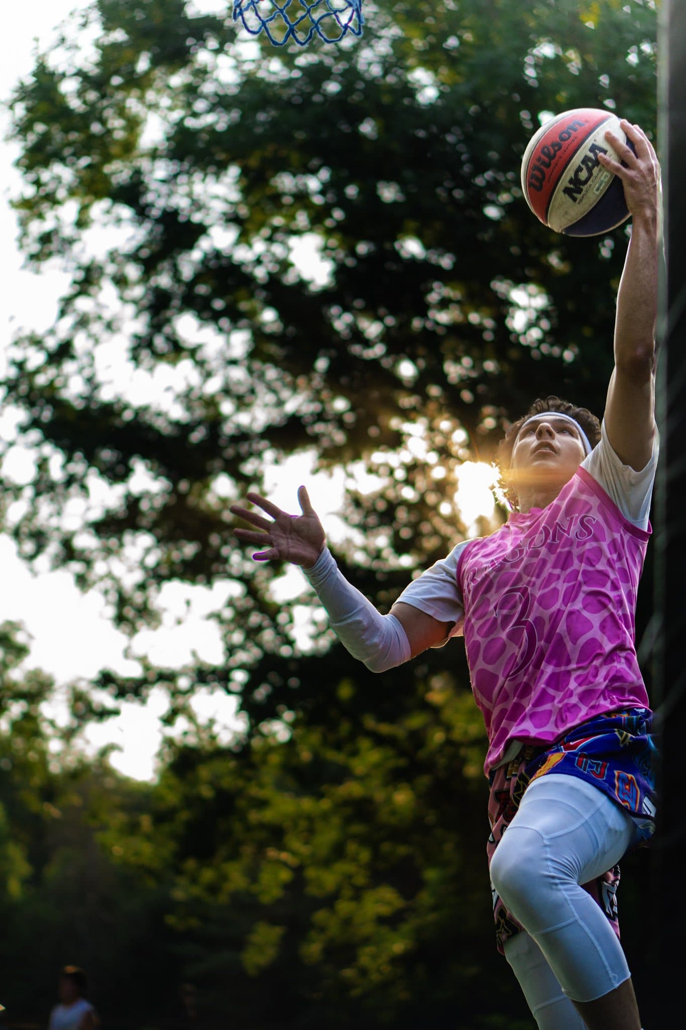 Player in a pink Dragons number 3 jersey going up for a layup, sun flaring through the trees