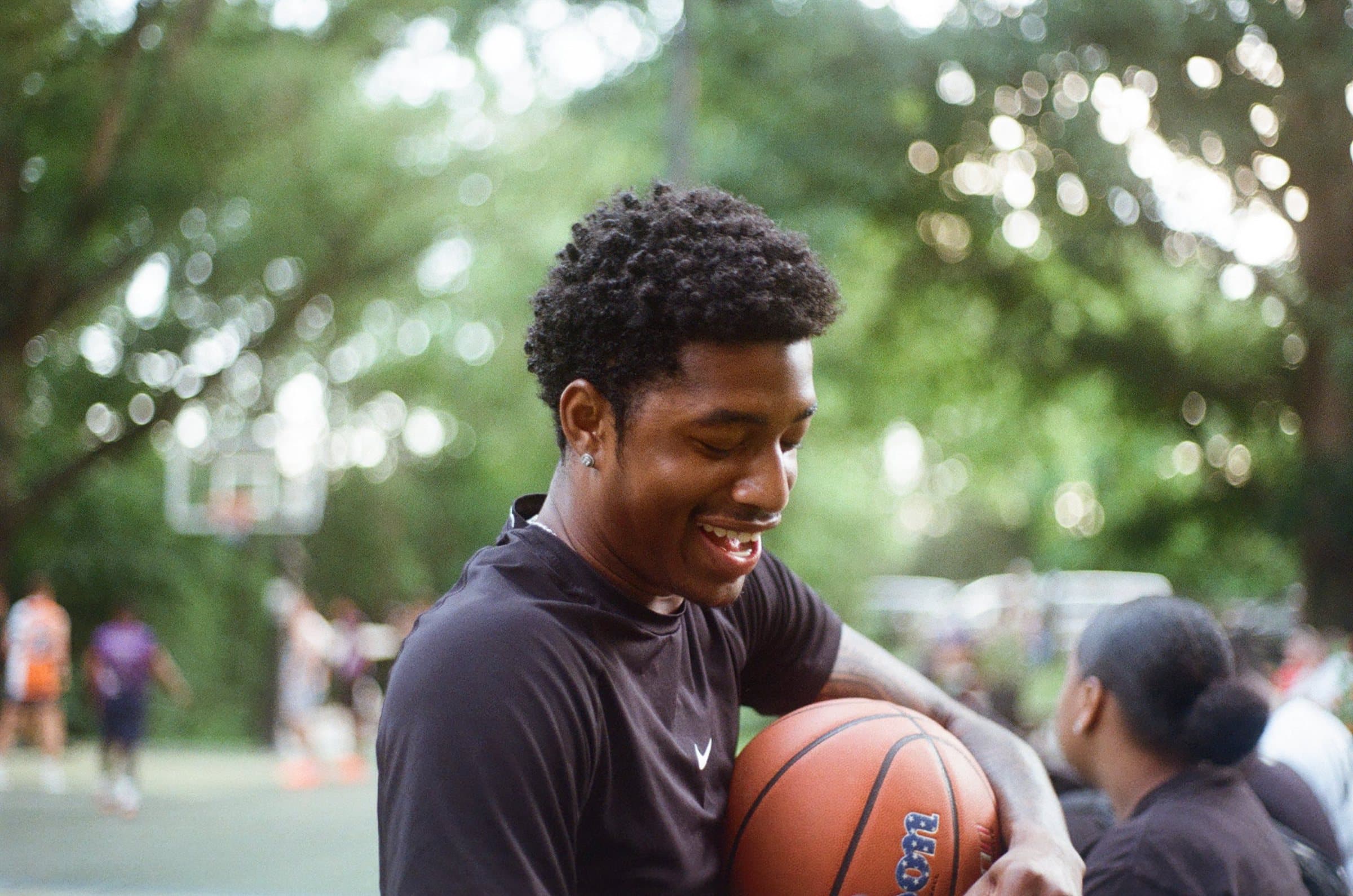 Player in a black t-shirt smiling down at the basketball in his hands, court and trees behind him