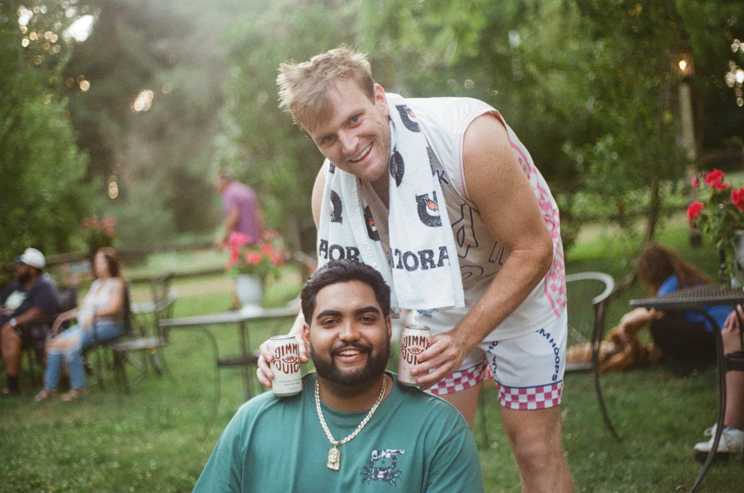 Two friends at a garden party, one holding Jimmy Juice cans up to the other's ears
