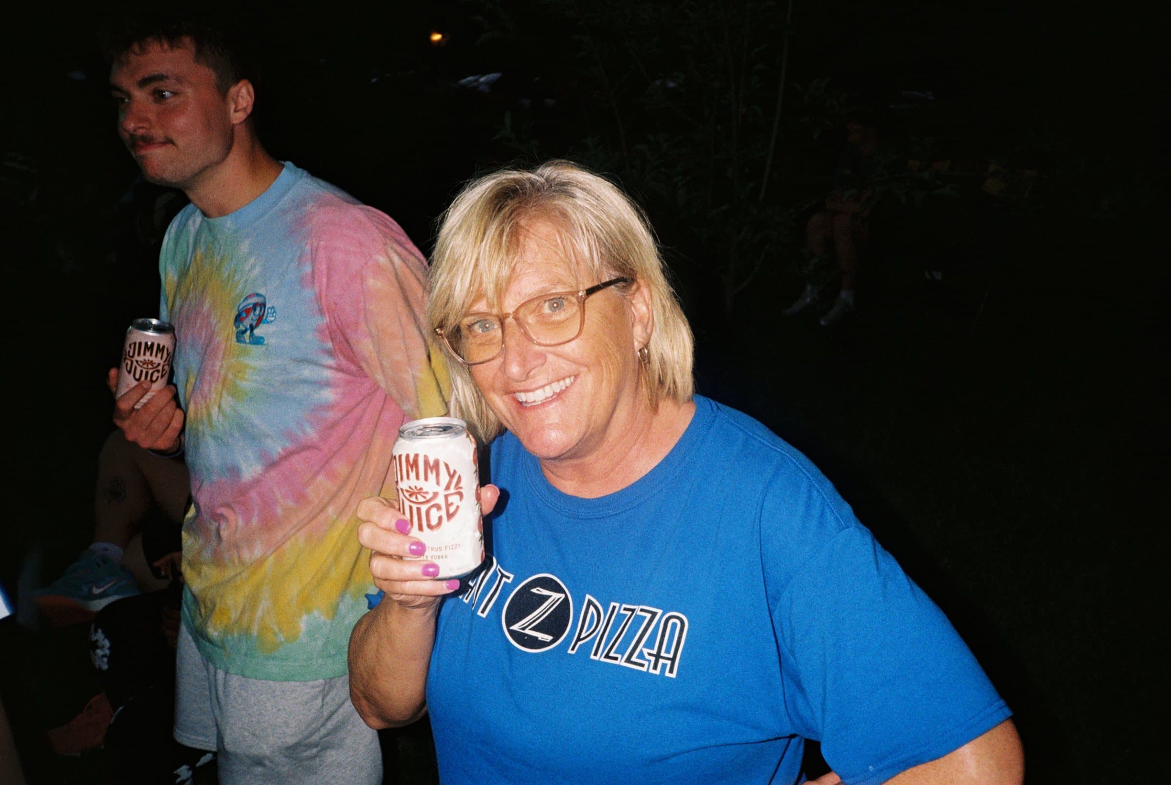 Woman in a blue t-shirt smiling and holding up a can of Jimmy Juice at night
