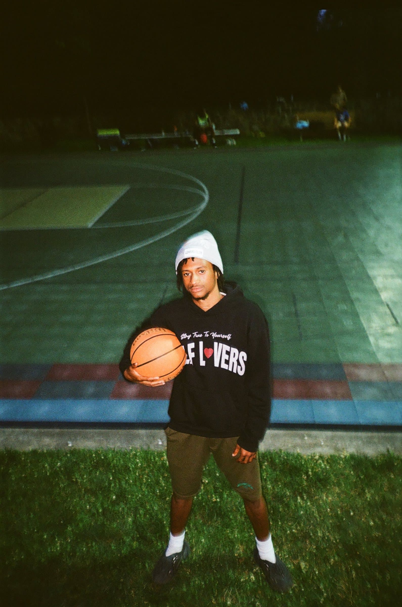 Young man in a black Self Lovers hoodie and white beanie holding a basketball beside the court at night
